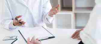 A healthcare professional in a white coat discusses with a patient who has a blood pressure cuff on their arm. There is a medical clipboard and a blood pressure monitor on the table between them. The background shows shelves out of focus.