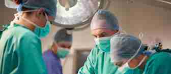 Three surgeons in green surgical gowns and masks perform an operation under a large surgical light. Another medical professional in purple scrubs stands in the background, observing. The setting appears to be a sterile operating room.