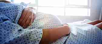 A pregnant person lying in a hospital bed wearing a hospital gown, with hands resting on their belly, sunlight streaming through a window in the background.