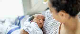 A newborn baby wrapped in a white blanket looks up at an adult holding them in a hospital bed. The adult wears a hospital gown, and the scene suggests a caring post-birth moment.