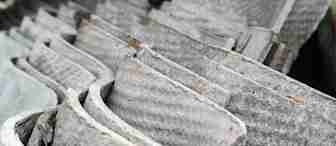 Stacked corrugated sheets of weathered, gray asbestos roofing material, showing a rough texture with visible fibers. The sheets are densely packed, illustrating an industrial or construction setting.