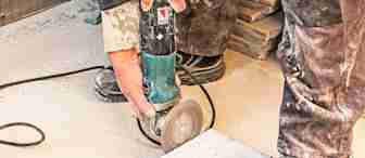 A person wearing dusty work clothes and sturdy shoes is using a power grinder to cut a piece of tile. The scene is dusty, suggesting active construction or renovation work. The tile is placed on top of another as the grinder partially cuts through it.