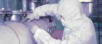 Person in full protective gear repairing a large industrial pipe. They are wearing a white hazmat suit, gloves, and a mask. The surroundings appear to be an industrial setting with various valves and pipes visible.