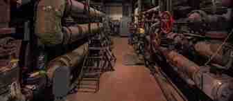 A dimly lit industrial room with aged, rusted pipes, and red valve wheels along the walls. Metal platforms and ladders are positioned around the equipment, with a few tools scattered on the floor. The atmosphere appears worn and gritty.

.