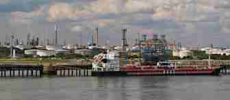 A large industrial port area with a docked cargo ship, surrounded by storage tanks and refinery structures. The water in the foreground is calm, and the background features numerous smokestacks and industrial buildings under a partly cloudy sky.
