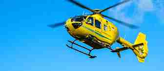 A yellow helicopter with green stripes flies across a clear blue sky. Its rotor blades are spinning, and there is a logo on the side. The helicopter is in motion, and the sunlight highlights its bright color.