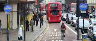 A busy city street with a dedicated bus and bicycle lane. A red double-decker bus is moving down the lane, followed by a bicyclist wearing a helmet and backpack. Pedestrians are walking on the sidewalk, and various vehicles are on the adjacent car lane.