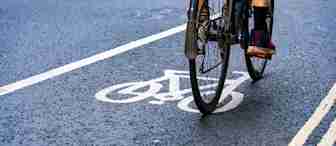 Close-up of a cyclist's legs and bike tires as they ride along a designated bike lane marked with a white bicycle symbol on an asphalt road. The person is wearing black socks and colorful sneakers. The surrounding road is slightly blurred, indicating motion.