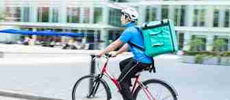 A delivery person rides a red bicycle on a city street, wearing a blue shirt, black pants, a white helmet, and carrying a large green insulated backpack. Blurred buildings and outdoor seating areas are visible in the background.