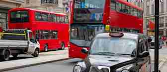 A busy urban street scene featuring two red double-decker buses, a traditional black taxi, and a service truck. The background showcases buildings adorned with British flags, indicating a vibrant city atmosphere.