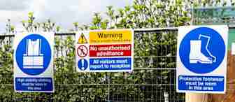 A metal fence displays multiple signs at a construction site. Signs include warnings for high visibility clothing, protective footwear, and no unauthorized admittance. There are also traffic cones and construction materials visible behind the fence.