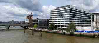 A large modern office building with glass windows stands on the banks of a wide river, under a cloudy sky. In the background, there's an older, towered structure with clock faces on each side. A bridge extends across the river on the left side.