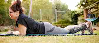 A person with glasses and hair in a bun is lying on a mat in a grassy area, supported on their elbows. They are wearing a black t-shirt, gray leggings, and sneakers. Their right leg is in a brace. Behind them, there is a green tree and a hammock.