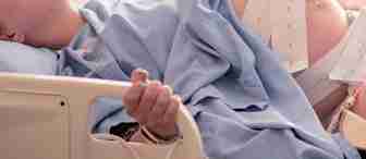 A pregnant person lies in a hospital bed, wearing a blue medical gown, with fetal monitoring straps around their exposed belly. Their eyes are closed, and one hand is gripping the bed railing. Medical monitoring equipment is partially visible beside the bed.
