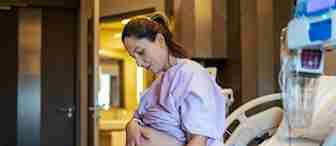 A pregnant woman in a hospital gown sits on a hospital bed while holding her belly with one hand. Medical equipment is visible next to the bed. Her expression looks focused and thoughtful. The room has a warm, softly lit atmosphere.