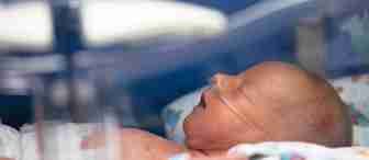 A newborn baby lies in an incubator. The baby has a nasal cannula providing oxygen and is swaddled with a patterned blanket. The image conveys a sense of care and medical support for the infant.