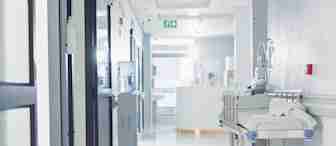 A hospital hallway with bright lighting. A medical gurney is parked against the wall, and a water cooler is nearby. In the background, a healthcare professional is visible at a reception desk. The corridor appears clean and sterile.