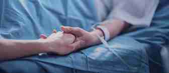 A close-up of two people holding hands. One is a patient lying in a hospital bed with an IV in their arm, while the other appears to be offering comfort and support. The scene conveys compassion and care in a medical setting.