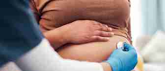 A healthcare professional wearing blue gloves and scrubs uses a stethoscope to listen to a pregnant person's belly. The pregnant individual is sitting and resting a hand on their abdomen, wearing a brown shirt and gray pants.
