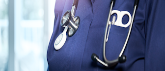 A close-up of a healthcare professional wearing a dark blue uniform with a stethoscope hanging around their neck. The background is blurred, indicating a clinical setting. The uniform features a pen in the chest pocket.