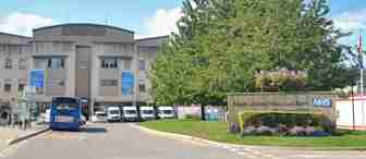 A hospital entrance with a sign reading "Royal United Hospitals Bath NHS." There’s a bus at a bus stop in front, several parked vans, and tall banners featuring a rainbow. A tree with flowers and some greenery are on the right side of the sign.