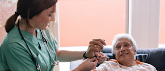 A nurse in a green uniform and stethoscope assists an elderly man in a striped shirt who is sitting on a couch near a window. The nurse is smiling while holding the man's wrist, checking his pulse or blood pressure. The man is relaxed and smiling back.