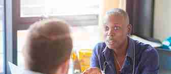 A healthcare professional with a stethoscope around their neck is talking to another person. The professional is gesturing with their hand while seated in a brightly lit office. The person they are speaking to is facing away from the camera.