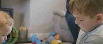 Two young children are playing with colorful toys on the floor of a living room. In the background, a woman sits on a couch, watching them. The room is softly lit, and a fireplace is visible on the left side.