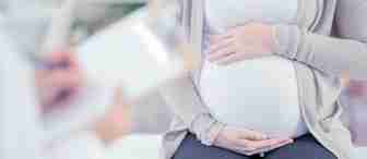 A pregnant person sits on an examination table, holding their belly. They are wearing a white shirt, a beige cardigan, and dark pants. In the foreground, a healthcare professional is taking notes on a clipboard. The setting appears to be a medical office.