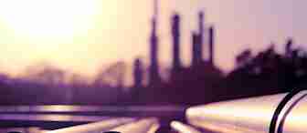 A close-up view of industrial pipelines extending towards the horizon. The background is blurred, revealing the silhouette of a refinery or factory under a vibrant sunrise or sunset sky, casting a purple-pink hue on the scene.
