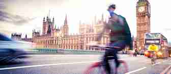 A blurred cyclist rides past the Houses of Parliament in London. The iconic Big Ben clock tower is clearly visible in the background. Nearby, a red double-decker bus and other vehicles are in motion, emphasizing the city's bustling atmosphere. The sky is partly cloudy.