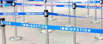 A zigzag queue setup with blue stanchions and barriers labeled "IMMIGRATION" in white text, typically found at an airport or border control area. The image shows an empty queuing space with multiple lines and posts.