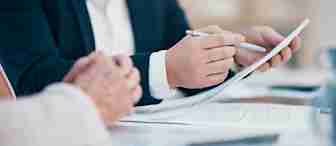 Two people are seated at a desk covered with paperwork. One person, partially visible and wearing a dark blazer, is holding a pen and pointing at a document. The other person's hands are visible, resting on the desk. The scene suggests a discussion or meeting.