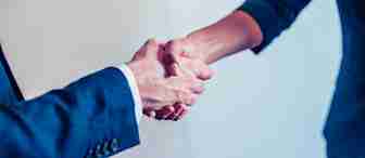 Close-up of two people shaking hands. One person is wearing a dark blue suit with a white shirt, while the other person is wearing a dark blue jacket. The background is blurred and light-colored, suggesting a professional or business setting.