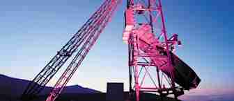 A striking image of a large antenna structure illuminated in pink and purple hues against a twilight sky with fading light. The silhouetted framework is set against a backdrop of distant hills, creating a visually captivating scene.
