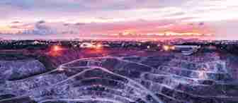A large open-pit mine with tiered levels, illuminated by several bright lights, stretches across the foreground under a colorful sky at sunset. In the distance, city lights twinkle, blending with the vibrant hues of the sky.