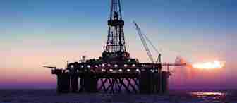 An offshore oil drilling platform is illuminated against a twilight sky over the ocean. A large flame is visible on the right side of the platform, suggesting gas flaring. A crane extends from the platform, and the sea is calm beneath.