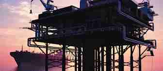 Silhouetted offshore oil platform in calm water during sunset, with a large ship anchored in the background. The sky is painted in hues of purple, pink, and orange, creating a serene and reflective atmosphere.