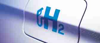 Close-up of a car's fuel cap cover with a blue "H₂" symbol, indicating it uses hydrogen as a fuel source. The silver metallic surface of the car is slightly blurred in the background.