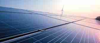 Close-up of solar panels with a wind turbine in the background under a clear sky. The sun is shining brightly, reflecting off the panels, representing renewable energy sources.
