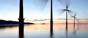 A row of wind turbines standing in calm water during sunset, with blurred blades indicating movement. The sky transitions from light blue to peach and pink near the horizon. Hills are faintly visible in the background.