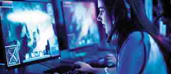 A person wearing a headset sits at a computer, engaged in a video game. The room is dimly lit with a blue hue. Multiple monitors display gameplay, featuring vibrant graphics. The person uses a keyboard and mouse, focused on the screen.