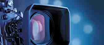 Close-up shot of a professional video camera lens with a large square hood, capturing a clear view of the lens glass. The background is blurred and has a gradient of blue tones with lights creating bokeh effects.