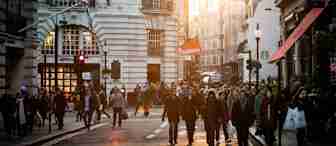 A busy city street at sunset with crowds of people crossing and walking along the sidewalks. Historic buildings with ornate facades line the street, and a warm golden light bathes the scene. British flags hang from the lamp posts, adding a sense of place.