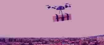 A drone is flying over a cityscape, carrying a package secured with black straps. The image has a pink overlay that tints the entire scene, including the buildings below and the sky above.
