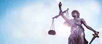 A bronze statue of Lady Justice holding scales in her left hand and a sword in her right hand, with a blindfold covering her eyes. The background is a clear, bright blue sky with light clouds. The image focuses on the statue, symbolizing fairness and law.