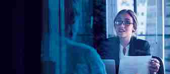 A woman wearing glasses and a business suit is holding a paper and speaking to another person across a desk in an office setting. The scene is viewed through a window, reflecting the cityscape outside.