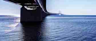 Photograph of a long bridge extending over a vast body of water, disappearing into the horizon. The bridge's underside is visible, with support pillars reflecting on the calm water. The scene is set under a clear blue sky during daylight.