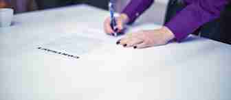 A person wearing a purple shirt is signing a contract on a white table with a pen. Another person in a blue suit stands opposite, with only their torso and hands visible. A small white coffee cup is placed on the table in the background.