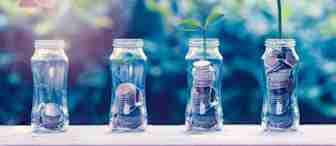 A hand places a coin into one of four glass jars filled with coins. Two jars have small plants sprouting from them, symbolizing financial growth and investment. The background is blurred greenery, highlighting the focus on the jars and coins.